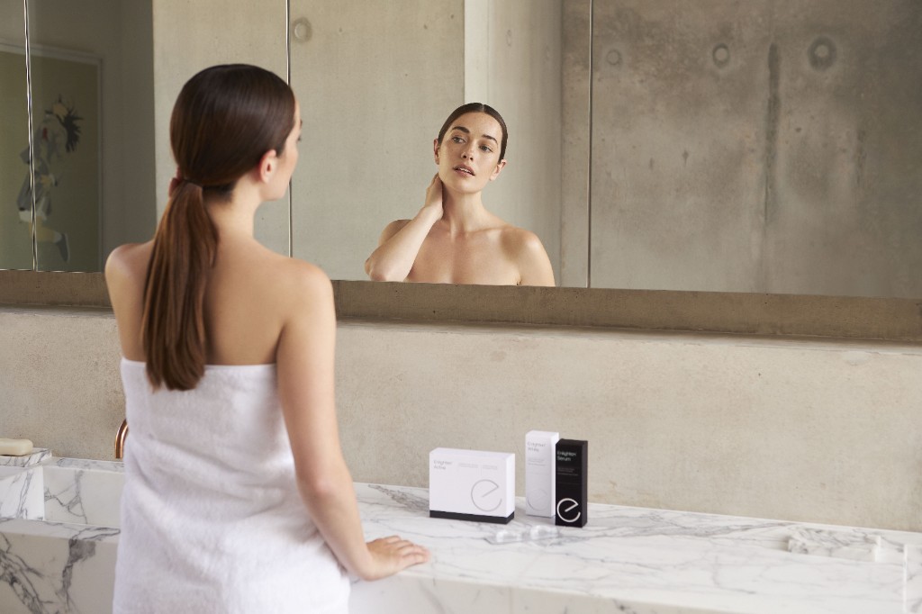 Donna allo specchio in bagno con i prodotti Enlighten Active, White e Serum